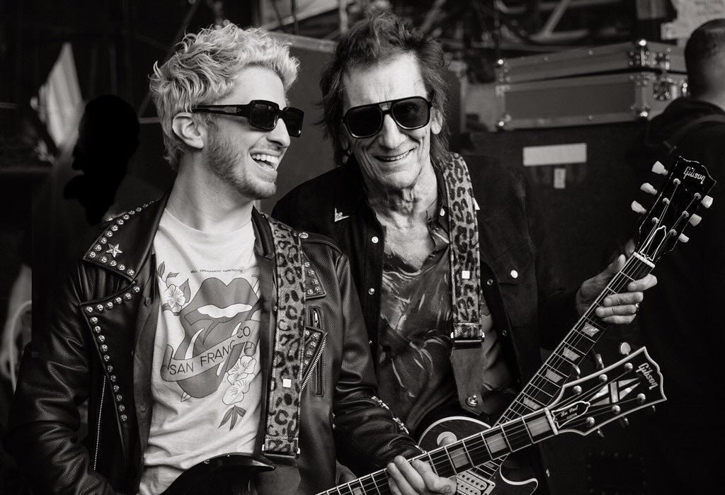 A black and white photograph of two rock musicians standing backstage, each holding a Gibson electric guitar. One wears a studded leather jacket and sunglasses, and the other wears a patterned leather jacket and also sunglasses.