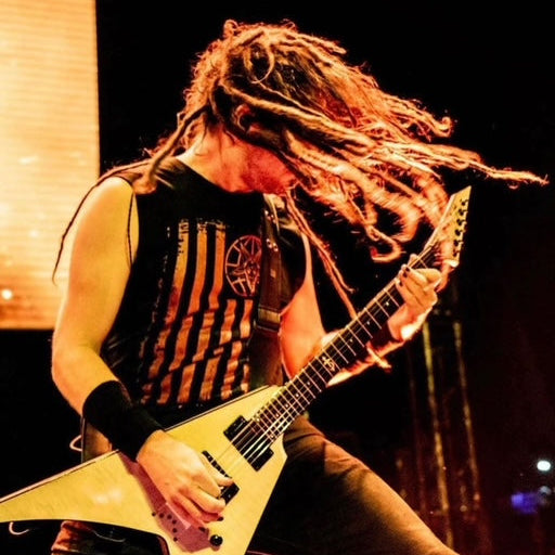 A rock musician with long dreadlocks playing a cream-colored angular electric guitar on stage, wearing a black sleeveless shirt with orange stripes.