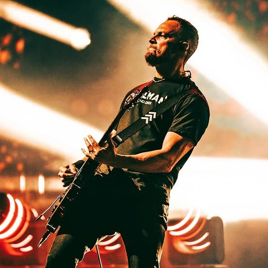 A male musician performing on stage, wearing a black graphic t-shirt, dark jeans, and white sneakers while holding an electric guitar under dramatic stage lighting.
