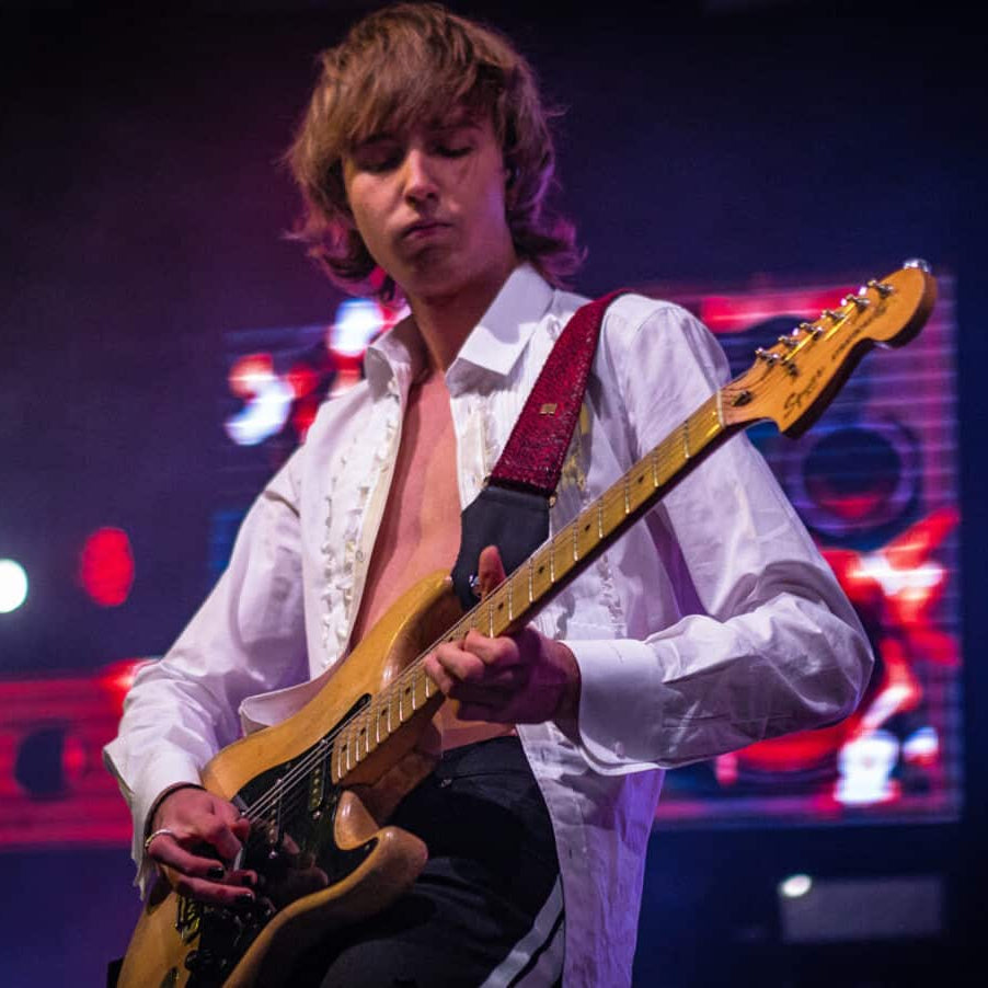 A male musician on stage playing a gold electric guitar, wearing a white open-collar shirt and dark trousers under dramatic red and blue stage lighting.